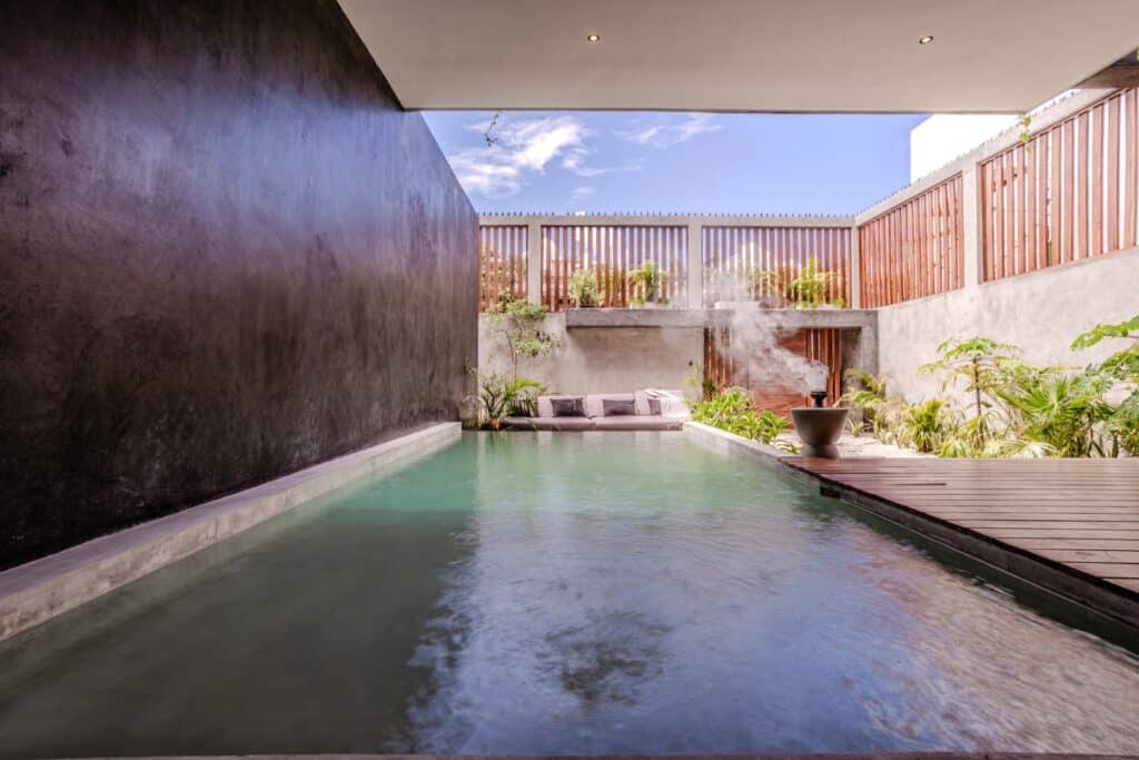 A modern outdoor pool area at Villa Karmond in Tulum's Region 15, featuring a steam feature, lush greenery, wooden fencing, and a cozy seating nook under a partially covered patio.