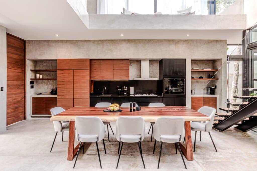 Modern kitchen and dining area at Villa Karmond in Tulum's Region 15, featuring wooden cabinets, black appliances, exposed concrete walls, and ample natural light.