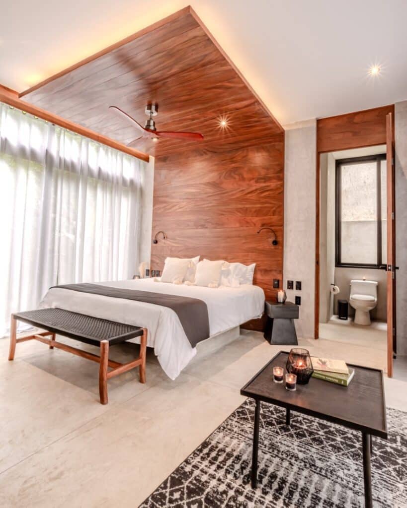 Modern bedroom in Villa Karmond, Tulum, featuring a wood accent wall, minimalist furniture, and a partial bathroom visible through an open door.
