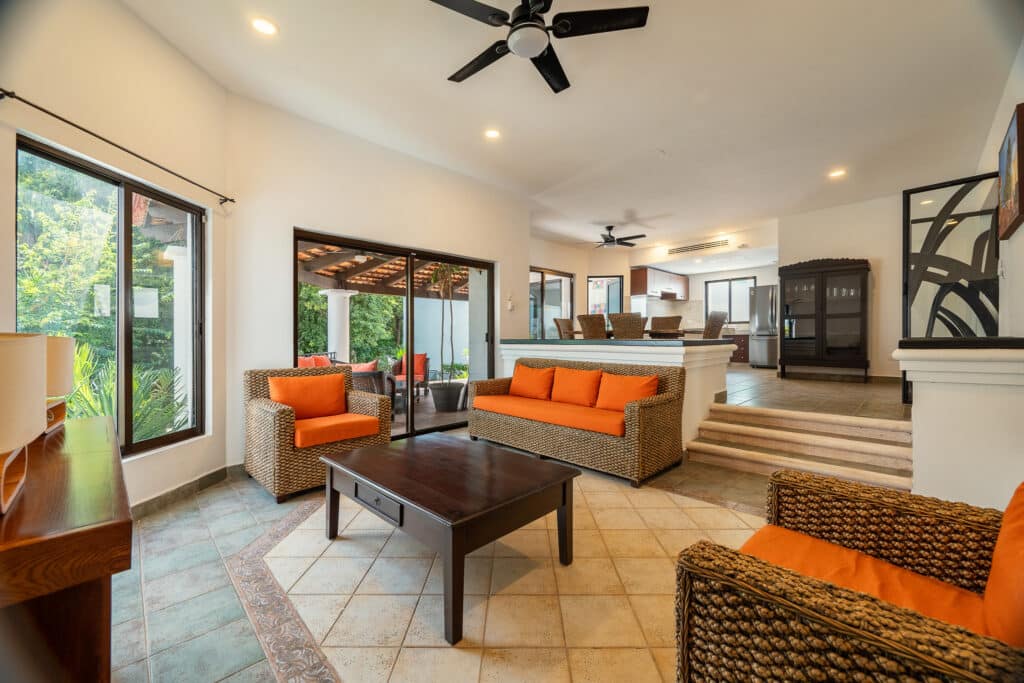 Interior view of Casa Marita in Playacar Phase 2, Playa del Carmen, featuring a living room with wicker furniture with orange cushions, tiled flooring, and large windows.