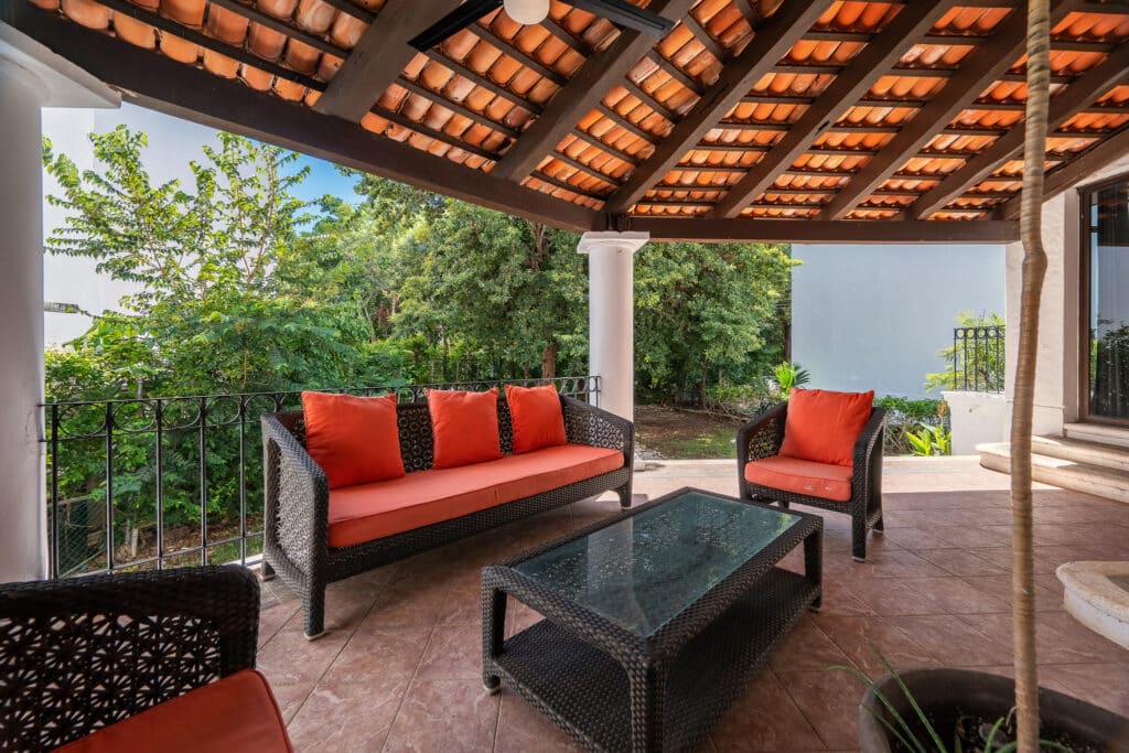 A covered patio at Casa Marita in Playacar Phase 2, Playa del Carmen, features black wicker seating with orange cushions, a glass-topped table, surrounded by lush greenery.