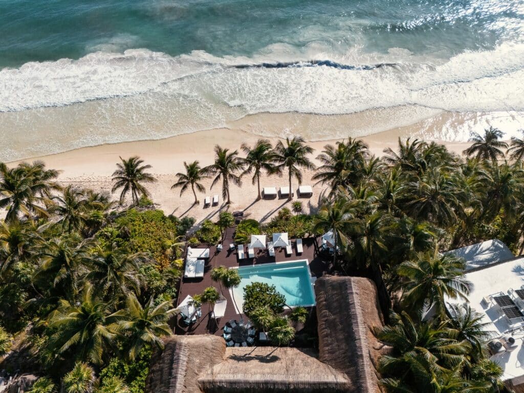 An aerial view of Casa Chic in Tulum showcasing a beachfront home with a pool surrounded by palm trees, outdoor seating, and direct access to the sandy shoreline.