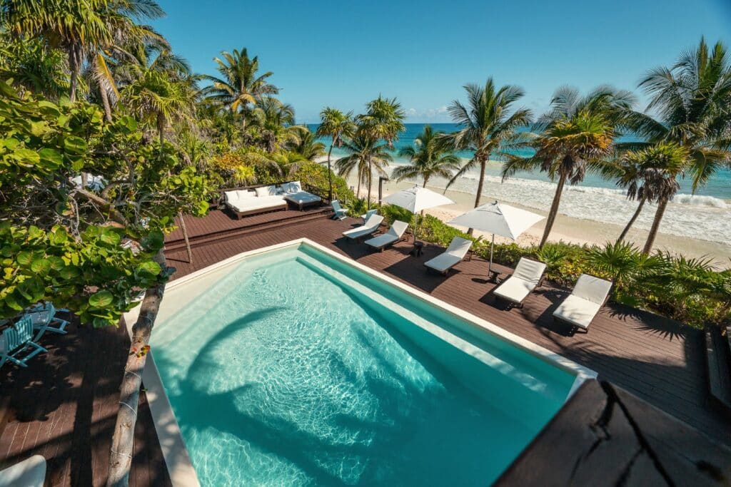 A beachfront home in Tulum features a private pool, surrounded by palm trees and lounge chairs overlooking the sandy shore and turquoise ocean.