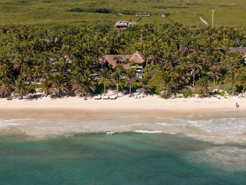 A beachfront home in Tulum surrounded by palm trees sits directly on the sandy shore with lounge chairs and a person walking along the water.