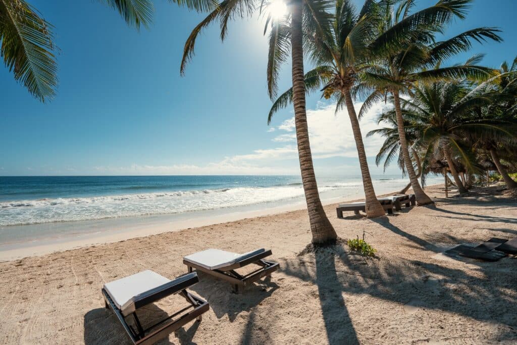 A beachfront home in Tulum, featuring palm trees, white sand, and lounge chairs under the sun with the ocean waves in the background.
