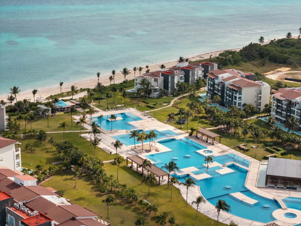 Aerial view of Mareazul in Playa del Carmen, showing a large pool area surrounded by modern condos, lush greenery, and nearby beach with turquoise water.