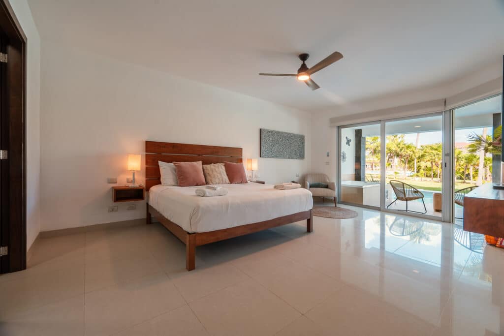 A spacious bedroom in Mareazul 401 Sur in Playa del Carmen, featuring a large bed with wood accents, a ceiling fan, and sliding glass doors leading to a balcony with a view of palm trees and a pool area.