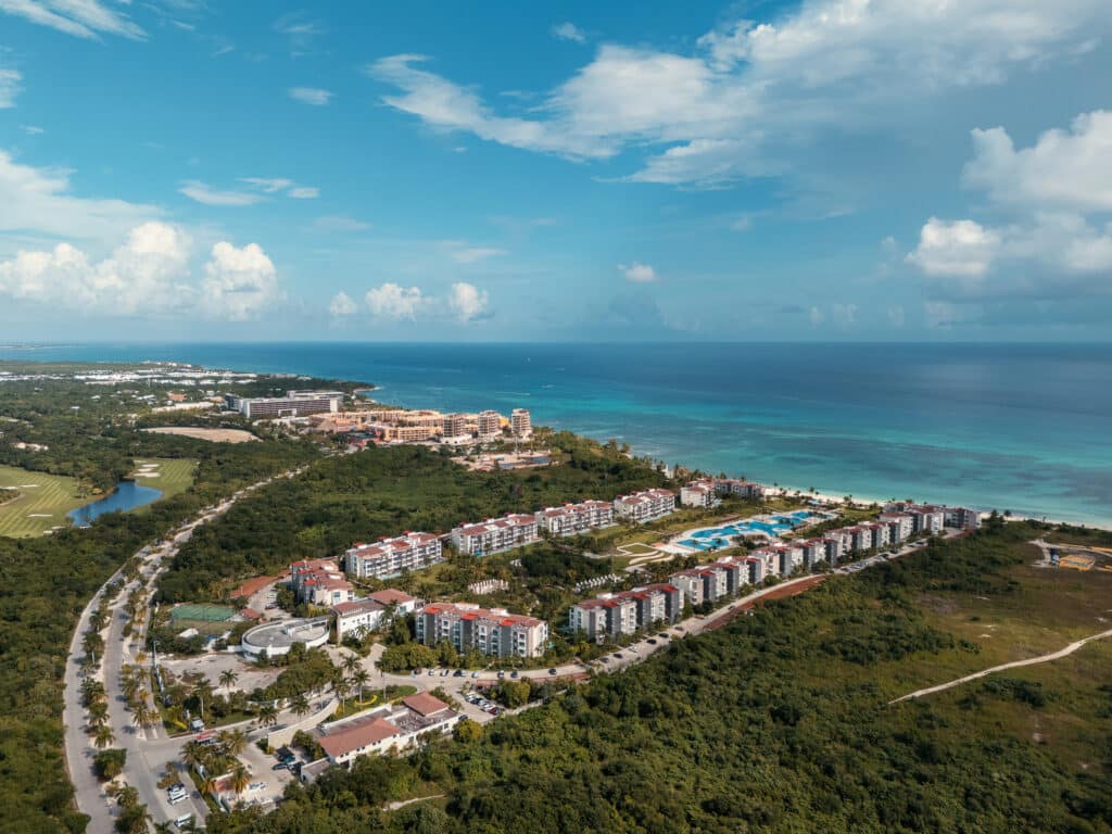 Aerial view of Mareazul in Corasol, Playa del Carmen, showing beachfront condo buildings, swimming pools, and lush greenery along the coast with blue skies above.