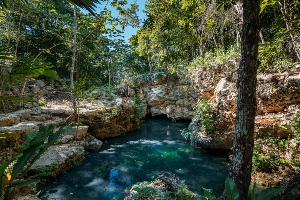 Two people sitting on rocks near a cenote with clear water, surrounded by lush green trees and rocky formations in Rancho San Martin, Akumal.