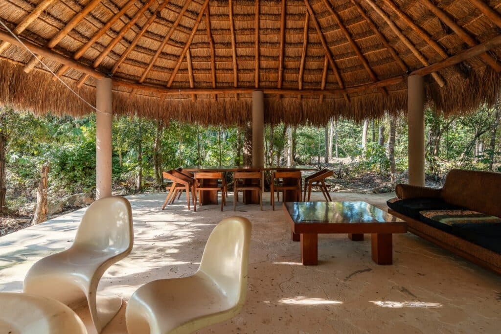 Open-air lounge with a thatched roof, wooden furniture, and modern white chairs, surrounded by lush green trees at Santuario in Akumal.