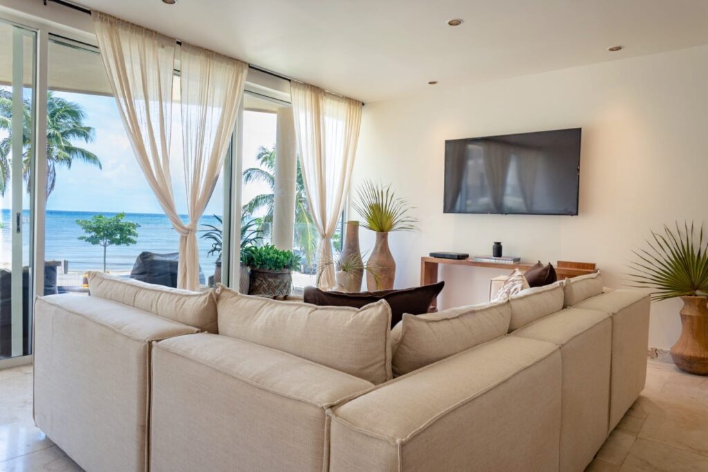 Living room at The Elements 124 in Playa del Carmen's Coco Beach area, featuring a beige sectional sofa, large windows with sheer curtains, and a view of the ocean.