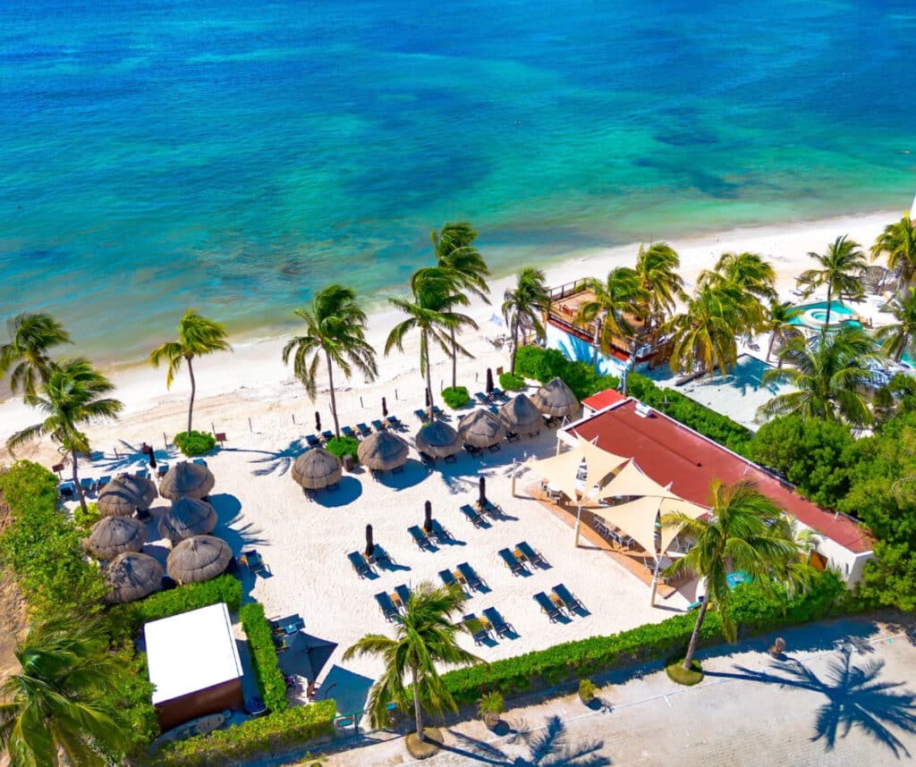 An aerial view of The Elements in Playa del Carmen's Coco Beach area, showing a beachfront pool area with sun loungers, shaded cabanas, and palm trees along the white sandy shoreline.