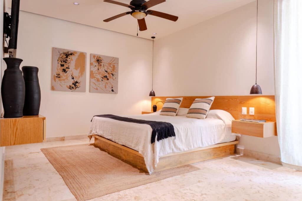 A modern bedroom in The Elements 124 development in Playa del Carmen's Coco Beach, featuring a wooden bed frame, neutral tones, and minimalist decor with artwork and hanging lights.
