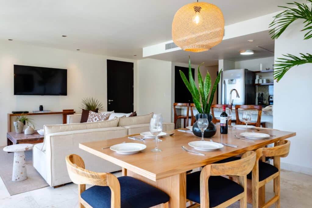 Interior of a condo at The Elements 124 in Playa del Carmen, featuring a open living and dining area with a wooden dining table, large plants, a mounted TV, and modern kitchen appliances.