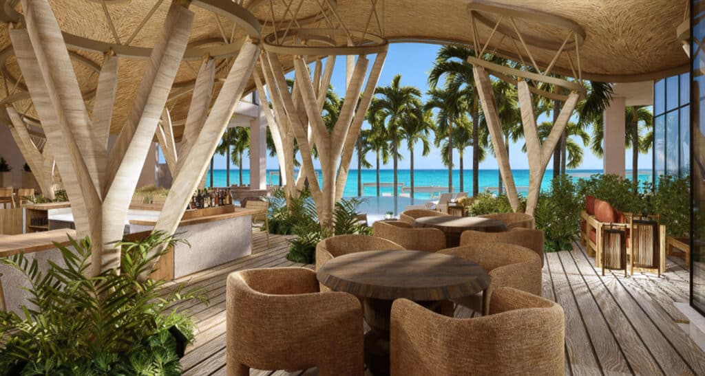 Interior view of Belmare 312 beachfront condo in Tulum, featuring a wooden ceiling, round tables with chairs, and large windows overlooking palm trees and the ocean.