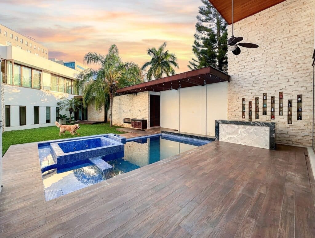A modern house in Bosques De Altabrisa, Merida, features a backyard with a small swimming pool, a grassy area with a dog, and a decorative wall with small shelves during sunset.
