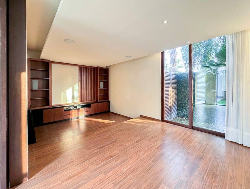 A spacious living area in Bosques De Altabrisa, Merida, featuring wooden flooring, built-in shelves, and large glass doors that open to an outdoor garden space.