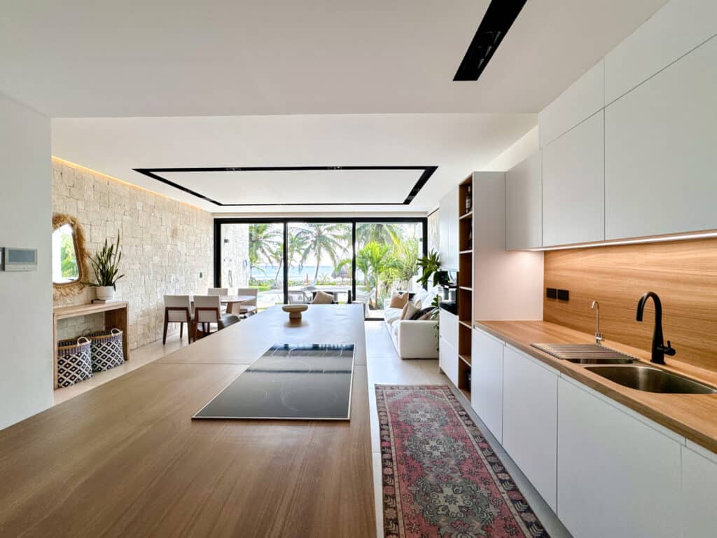 Interior view of a modern beachfront condo at CasaCún in Puerto Aventuras, featuring a sleek kitchen with wooden accents, a dining area, and a living space opening to outdoor palm views.