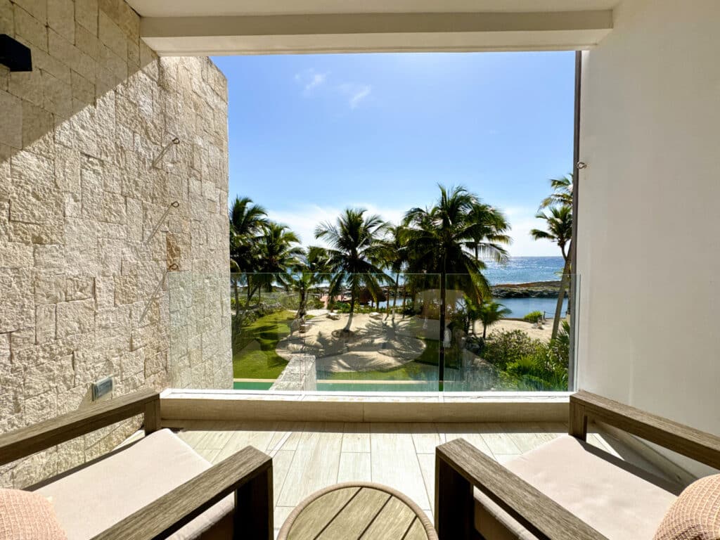 View from a beachfront condo at CasaCún in Puerto Aventuras showing a balcony with chairs, palm trees, a sandy area, and the ocean in the background.