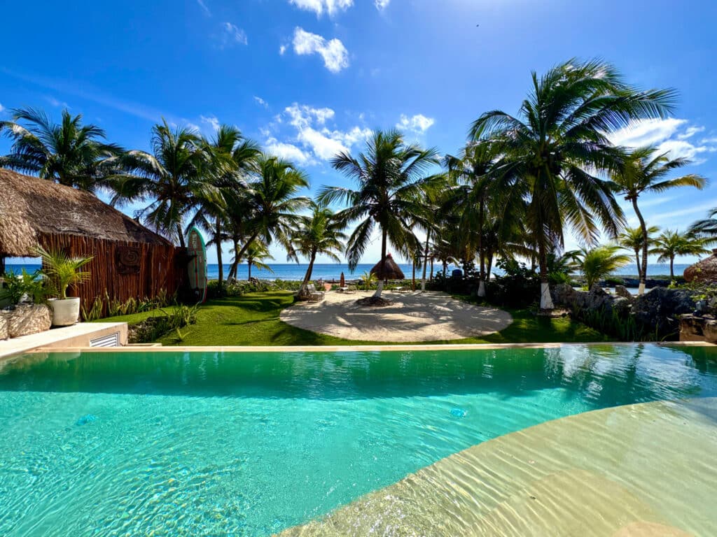 A beachfront view from CasaCún in Puerto Aventuras shows a swimming pool in the foreground, surrounded by palm trees, a sandy area, and the ocean under a blue sky.