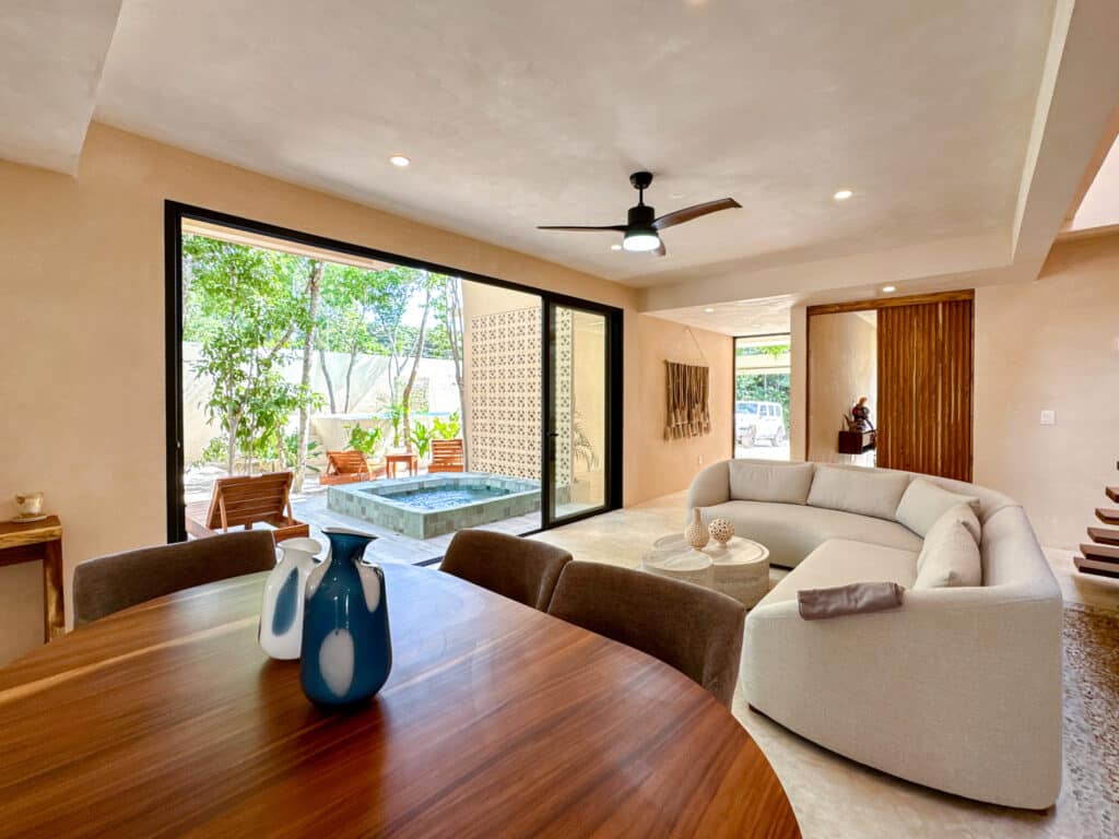 Living room of Casa Organica in Tulum featuring a curved sofa, a wooden dining table with vases, and a view of the outdoor area with a small pool and lush greenery.
