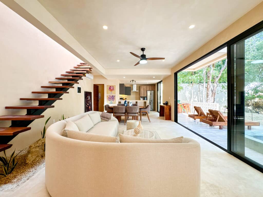 Open-concept living room in Casa Organica, Tulum, featuring a curved cream sofa, modern staircase, and large sliding glass doors leading to a shaded outdoor terrace surrounded by trees.