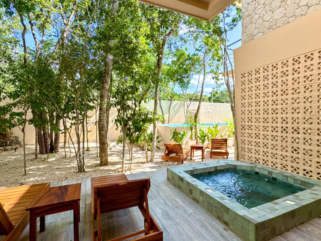 A shaded outdoor patio at Casa Organica in Tulum features a small tiled hot tub, wooden lounge chairs, and lush trees providing privacy and a natural serene atmosphere.