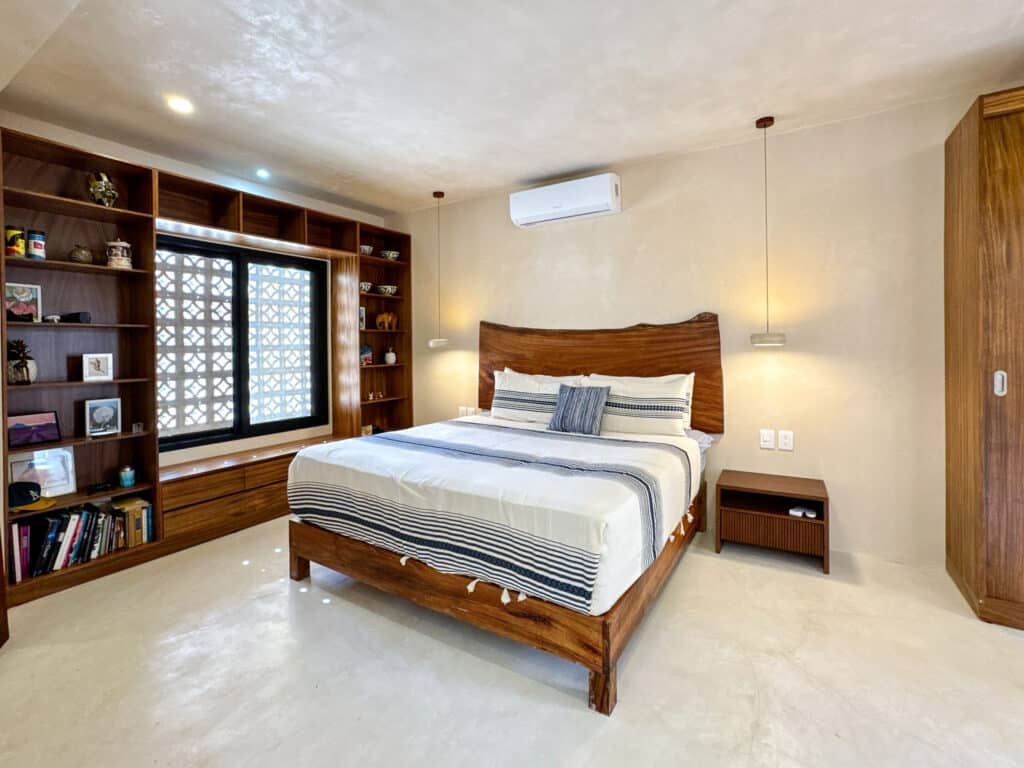 A spacious bedroom in Casa Organica with a large wooden bed, built-in bookshelves, and natural light coming through a window in Tulum's Region 15.