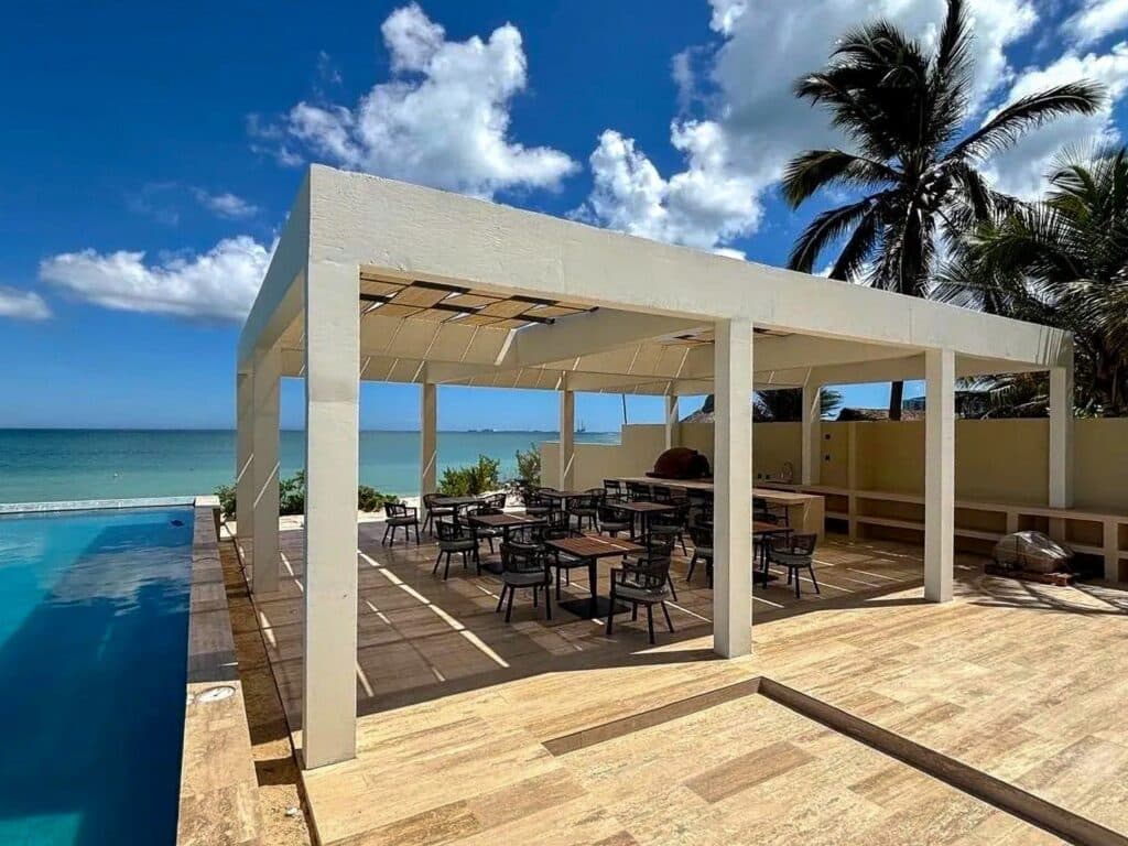 A covered terrace with tables and chairs overlooking the beachfront in Progreso, part of the Catavento A development, with a swimming pool and palm trees under a blue sky.