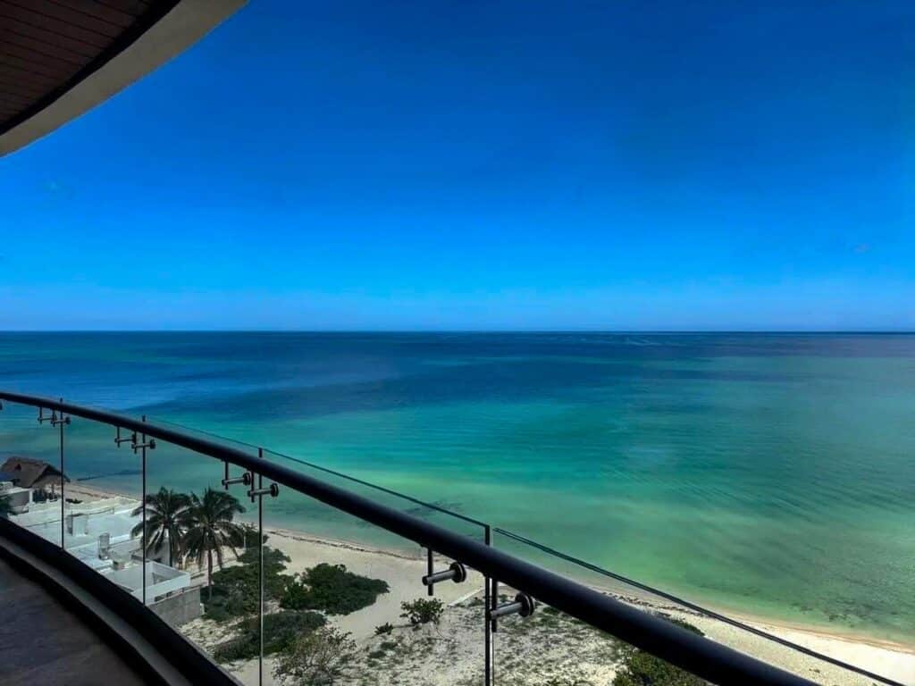 View from Catavento A-41 condo in Progreso Beachfront showing a clear blue sky, calm turquoise waters, and a sandy beach with palm trees.