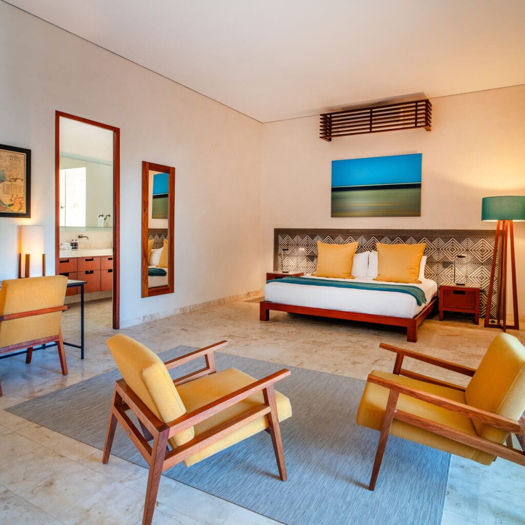 Interior of the El Pueblo Merida hotel in Centro Merida, featuring a spacious bedroom with a king-sized bed, yellow chairs, and modern decor.