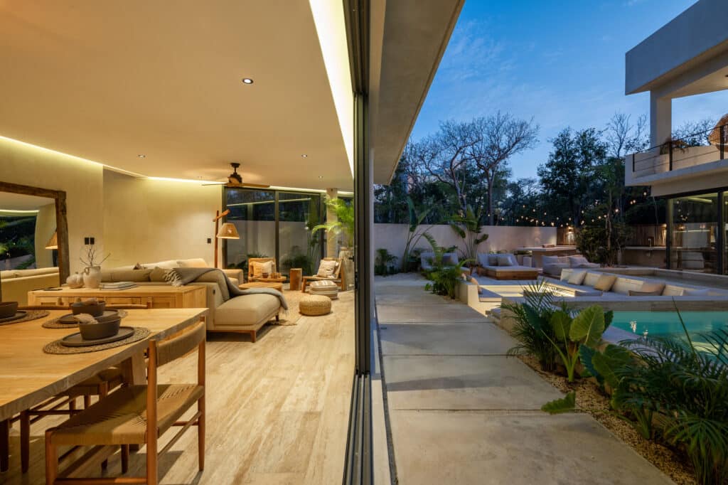 Interior and exterior view of Twolum in Tulum, showcasing a spacious living area with neutral decor, and a backyard with a pool, lounge seating, and lush greenery.
