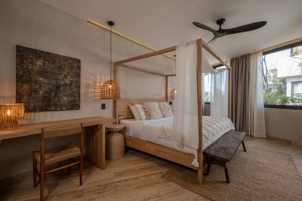 A bedroom in Twolum in Tulum features a wooden four-poster bed with sheer curtains, a rustic bench at the foot, woven pendant lighting, and natural tones throughout.