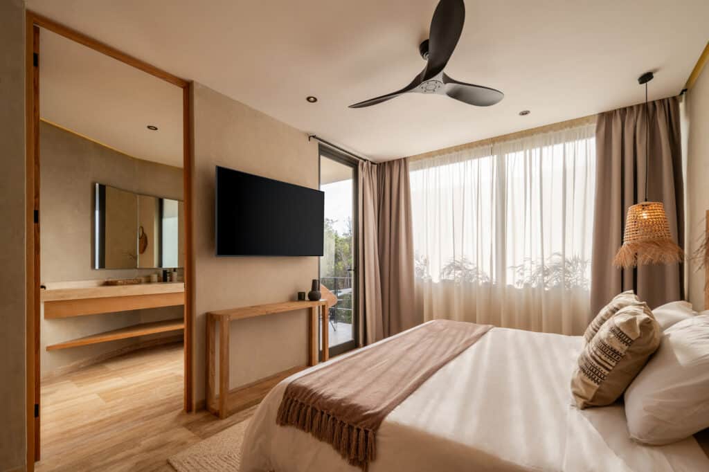 Interior view of a bedroom at Twolum in Tulum, featuring neutral tones, a ceiling fan, a mounted TV, a large window with curtains, and minimalist decor.