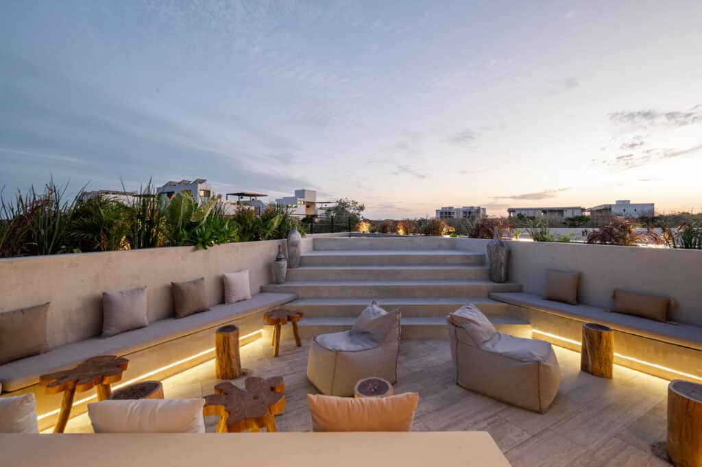 A rooftop terrace at Twolum in Tulum features built-in seating with cushions, small wooden tables, and lush plants against a sunset sky backdrop.