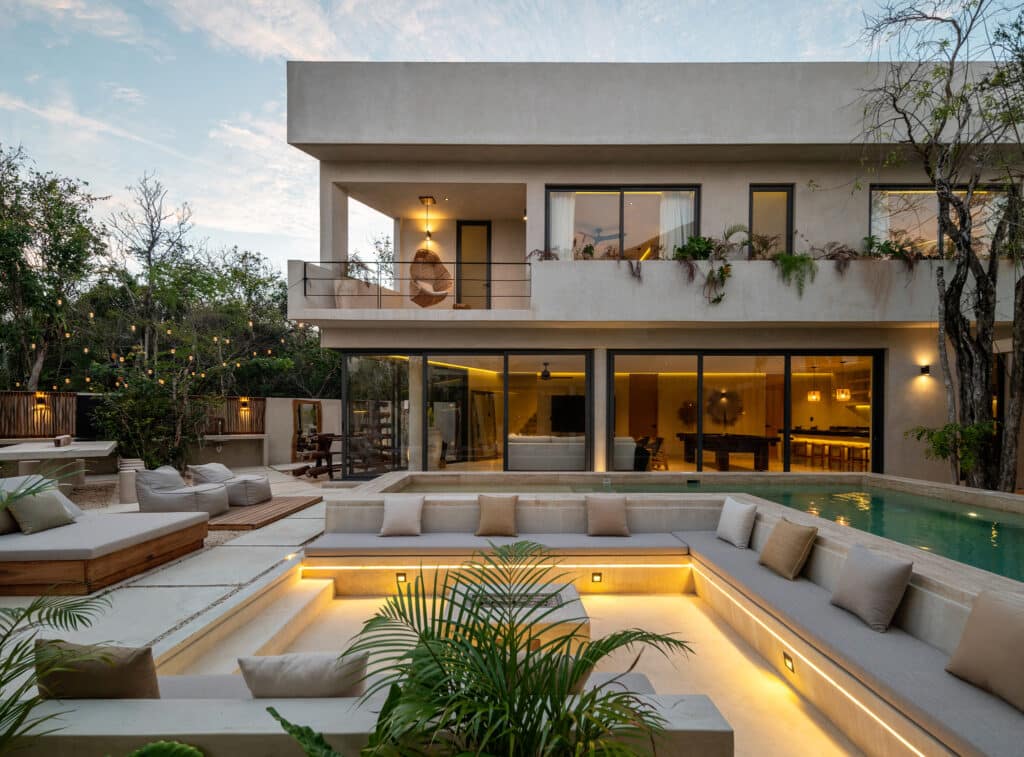 A modern villa in Tulum, features a spacious outdoor area with a pool, lounge seating, and a balcony with a hanging chair, set against lush greenery at dusk.