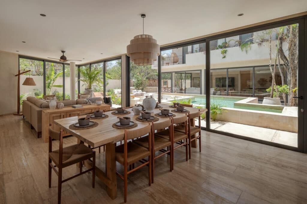 Interior view of Twolum in Tulum, featuring a spacious dining area with a large wooden table, overlooking a pool and lush outdoor garden through floor-to-ceiling glass doors.