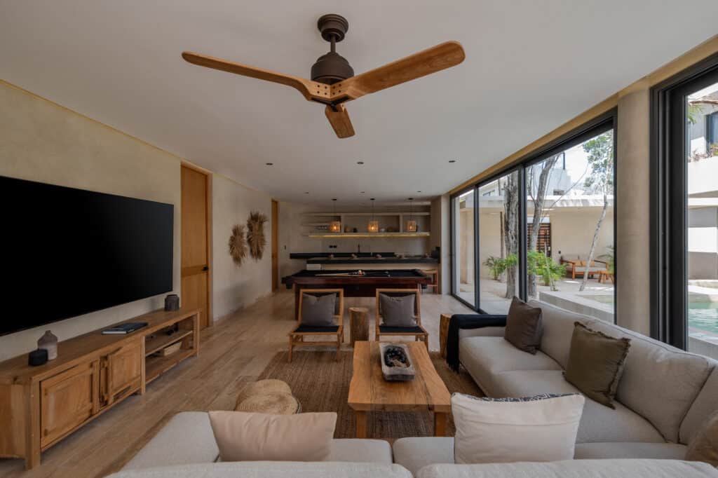 A spacious living room in Twolum, Tulum, featuring a beige sofa, wooden furniture, a ceiling fan, large glass doors opening to an outdoor area with trees.
