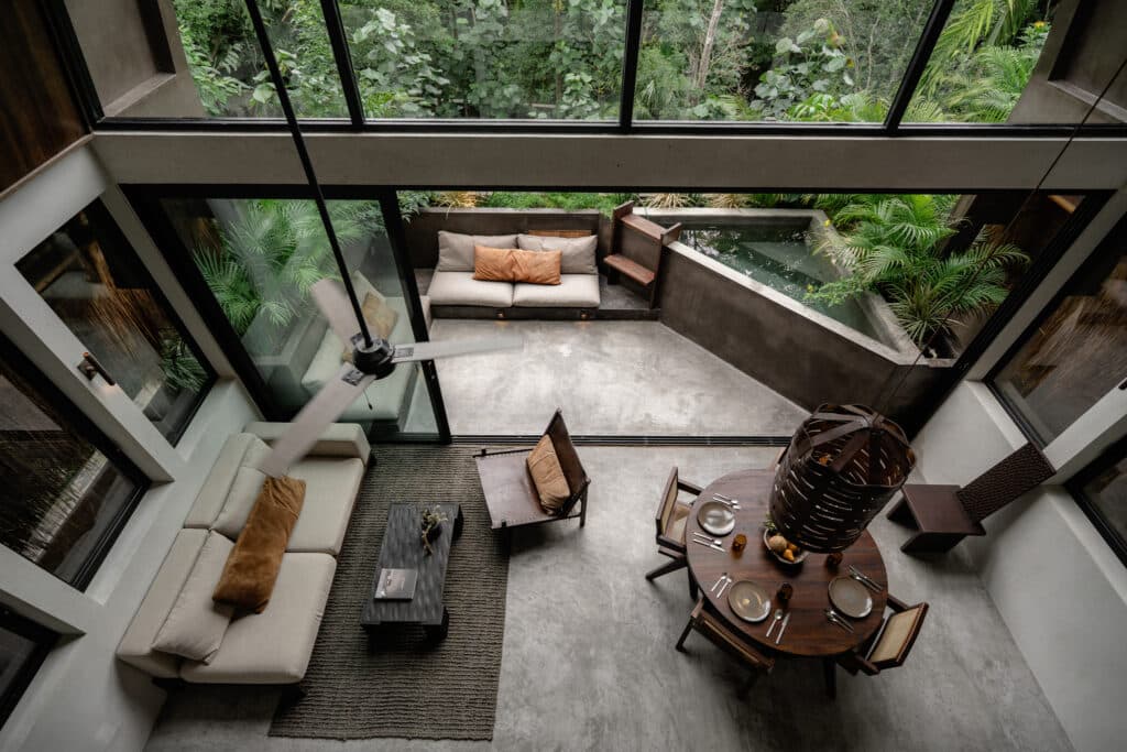 A spacious living and dining area with large glass windows overlooking lush greenery in Casa Kuro 203 in Tulum, featuring modern furniture and a high ceiling fan.