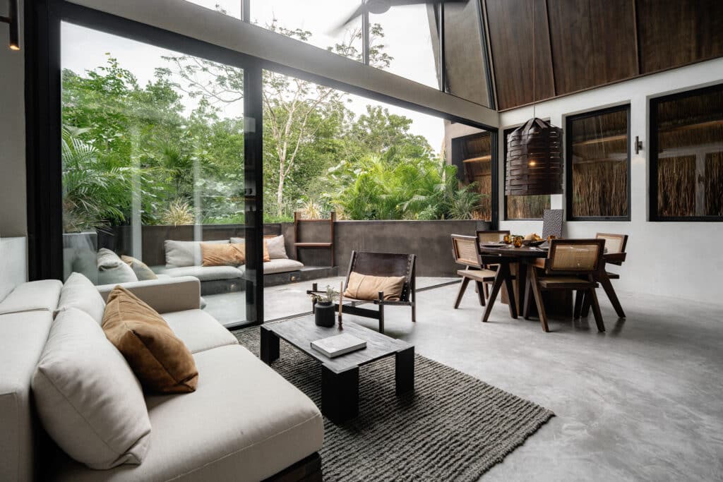 A spacious living area in Casa Kuro 203 in Tulum features large glass doors opening to a lush tropical patio, with contemporary furniture and natural wooden accents.