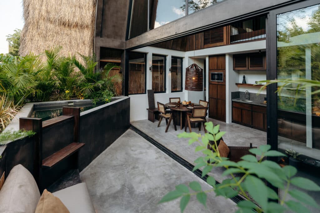 A modern outdoor dining area at Casa Kuro 203 in Tulum features a round wooden table, chairs, lush greenery, and large glass windows showcasing a contemporary design.