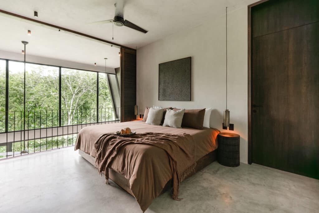 A modern bedroom in Casa Kuro 203 in Tulum features a minimalist design with a large bed, neutral tones, a dark wood door, and a balcony overlooking lush greenery.