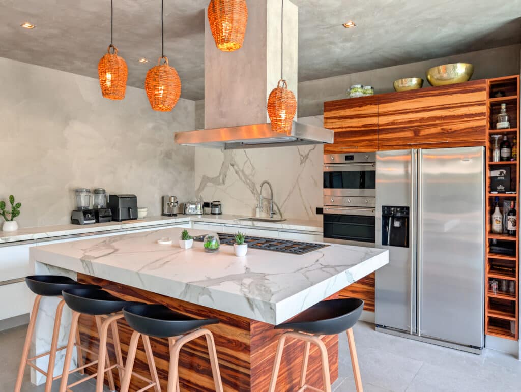 A modern kitchen in Casa Luum House, Playa del Carmen, featuring a marble island, wooden cabinetry, stainless steel appliances, and woven pendant lights.