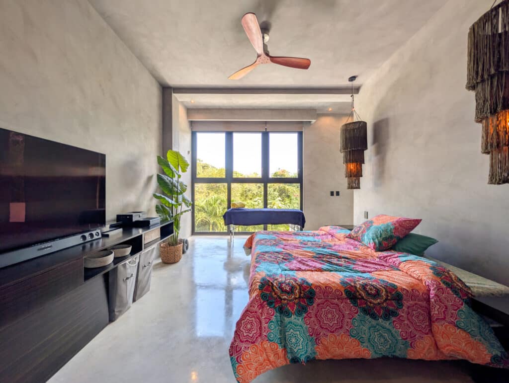 Interior view of Casa Luum House in Playacar Phase 2, Playa del Carmen, featuring a modern concrete design, a large window with greenery outside, a ceiling fan, and vibrant bedding.