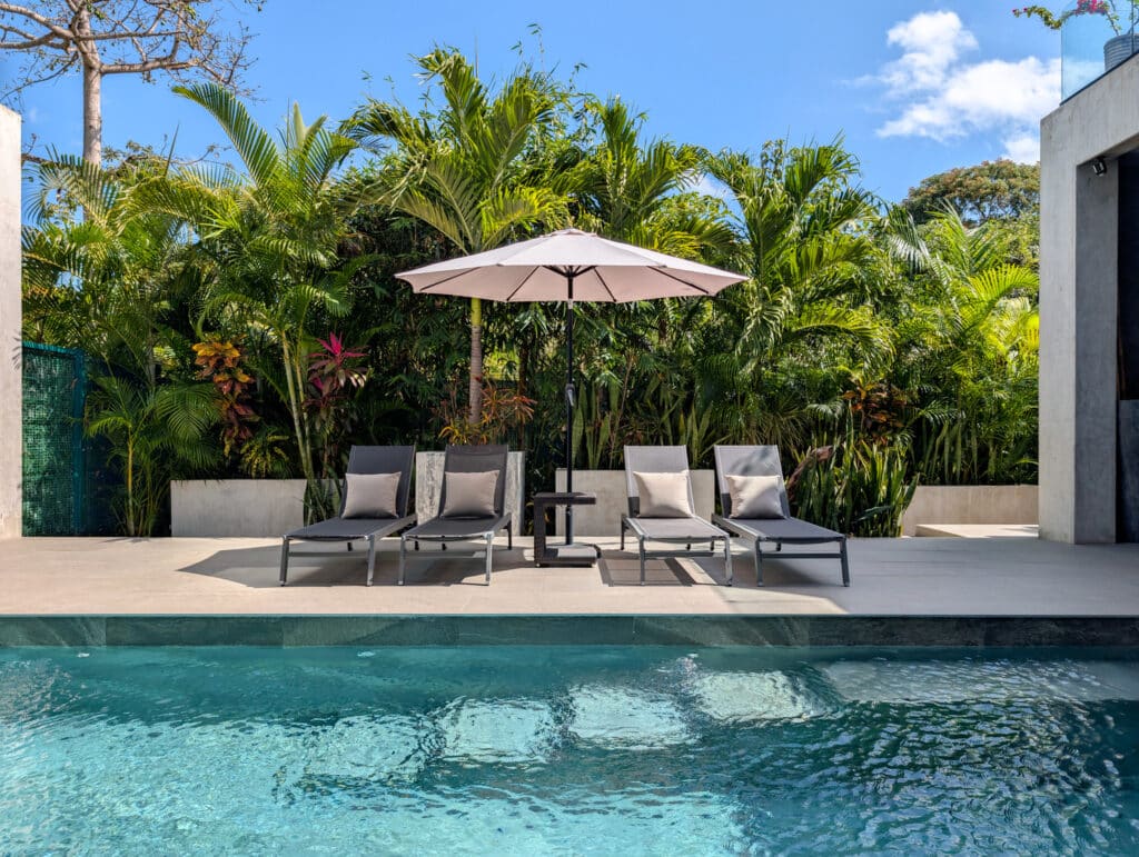 A view of the outdoor area at Casa Luum House in Playacar Phase 2, Playa del Carmen, featuring four lounge chairs with pillows, a large umbrella, and lush tropical foliage behind.
