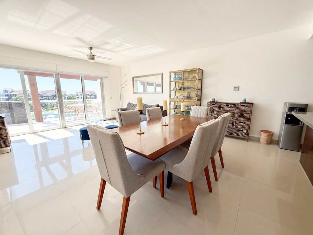 Dining area with a wooden table and light-colored chairs, adjacent to sliding glass doors leading to a balcony with outdoor furniture and a view of the pool at Mareazul 332N in Playa del Carmen.