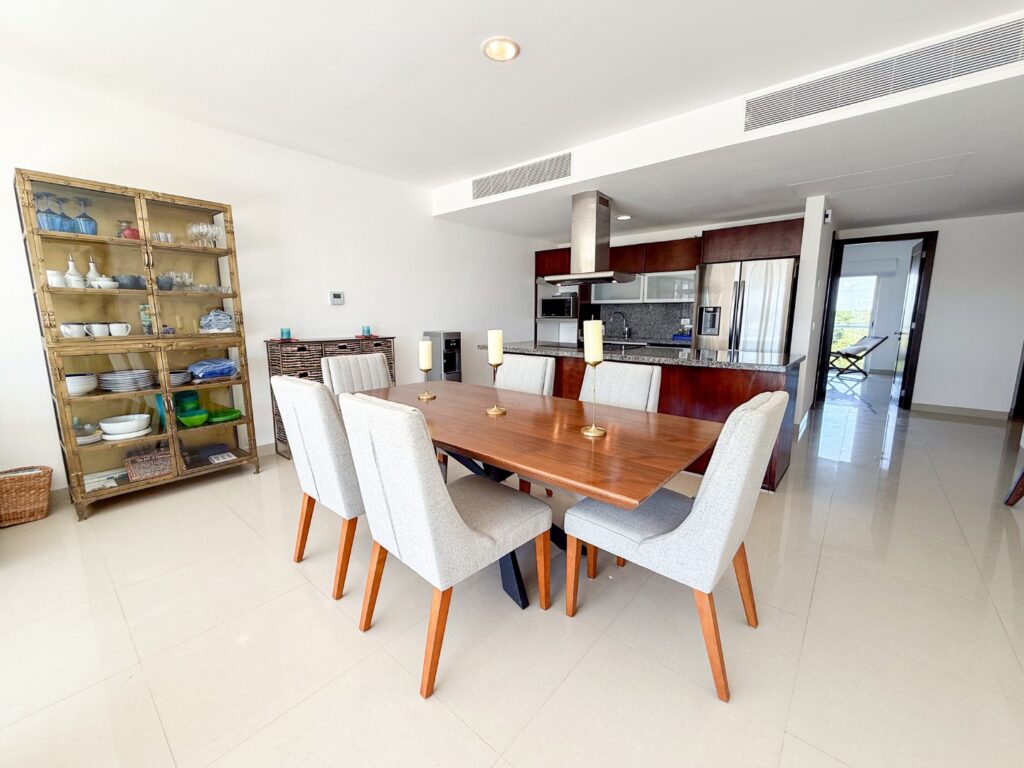 Interior view of Mareazul 332N penthouse in Corasol, Playa del Carmen, showing a dining area with a wooden table, light-colored chairs, and a kitchen with dark wood cabinets and stainless steel appliances.