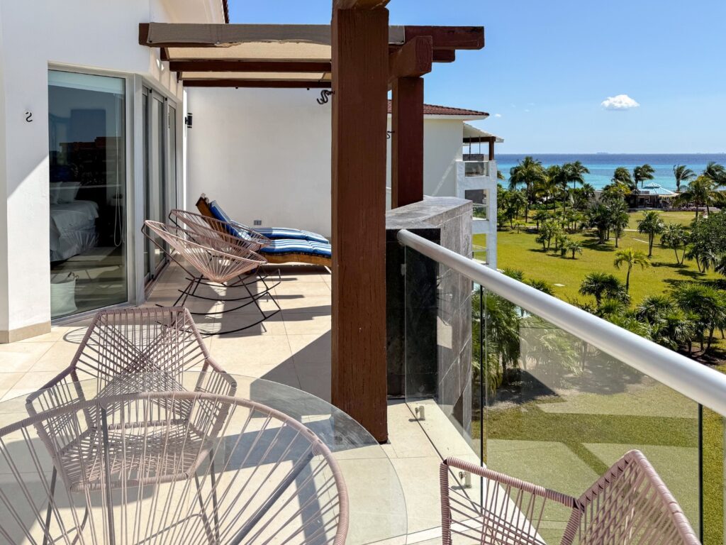 Balcony view from Mareazul 332N in Corasol, Playa del Carmen, showing outdoor furniture, lush greenery, palm trees, and the ocean in the background.