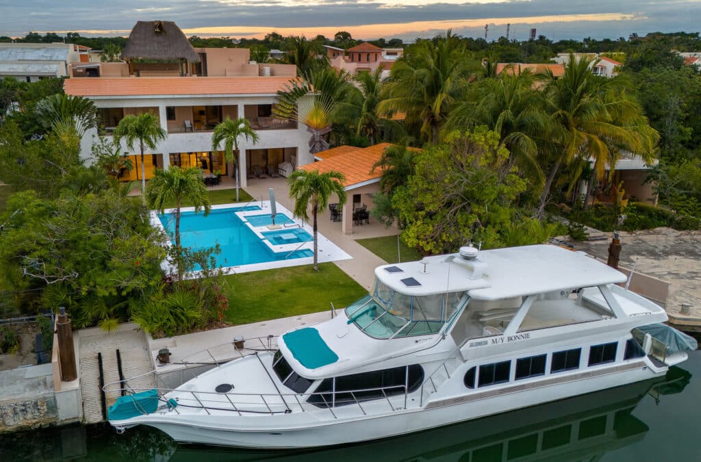 Marina Palms in Puerto Aventuras features a marina front property with a large white yacht, lush green palm trees, a swimming pool, and a modern building with terracotta roof tiles.