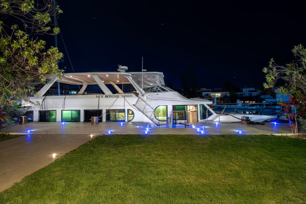 Night view of a large white yacht docked at Marina Palms in Puerto Aventuras, with green-lit windows, a satellite system, and illuminated deck, surrounded by greenery.
